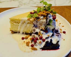 New York Cheesecacke mit Heidelbeerengrütze, Spekulatiuskekskrümeln und Himbeer-Schoko-Perlen auf einem Vanillesoßenspiegel.